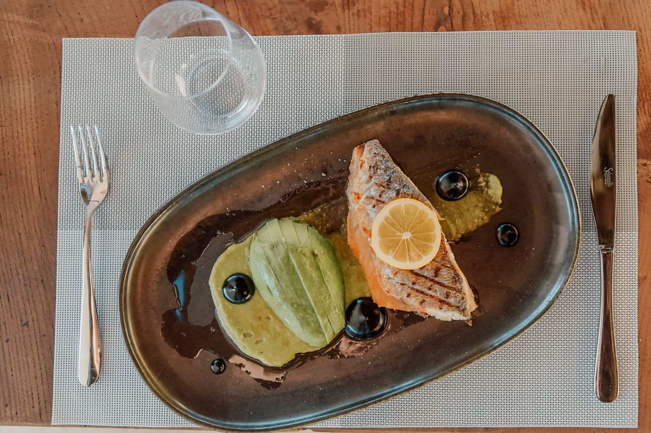 Close-up of a salmon dish with avocado and olives, elegantly plated on a gray tray with lemon slice.